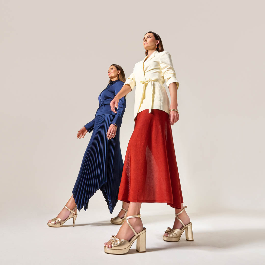 Two women wearing stylish outfits and daisy champagne heels with a plain background