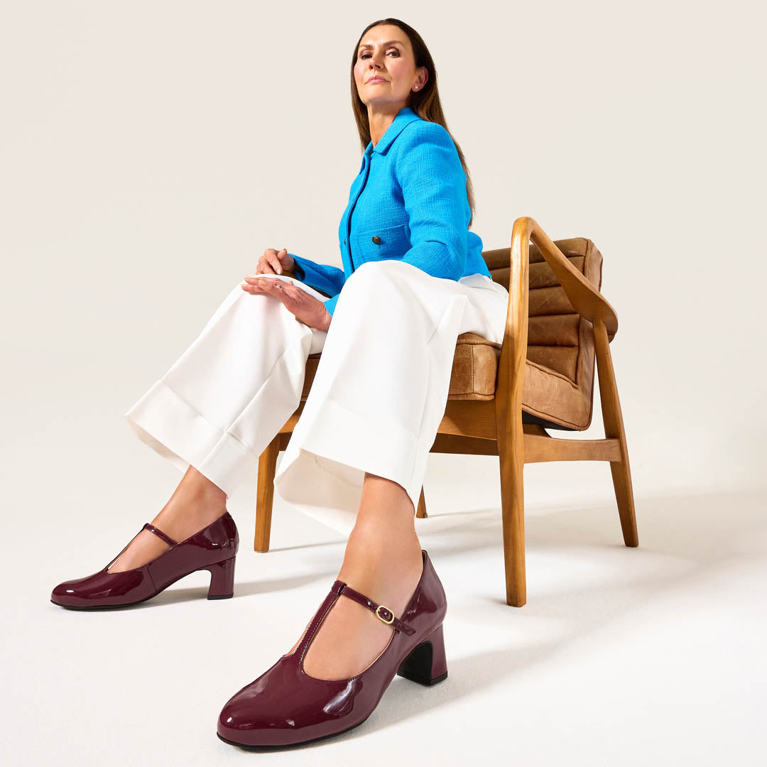 Woman sitting on a chair wearing a blue blazer, white pants, and maroon shoes.
