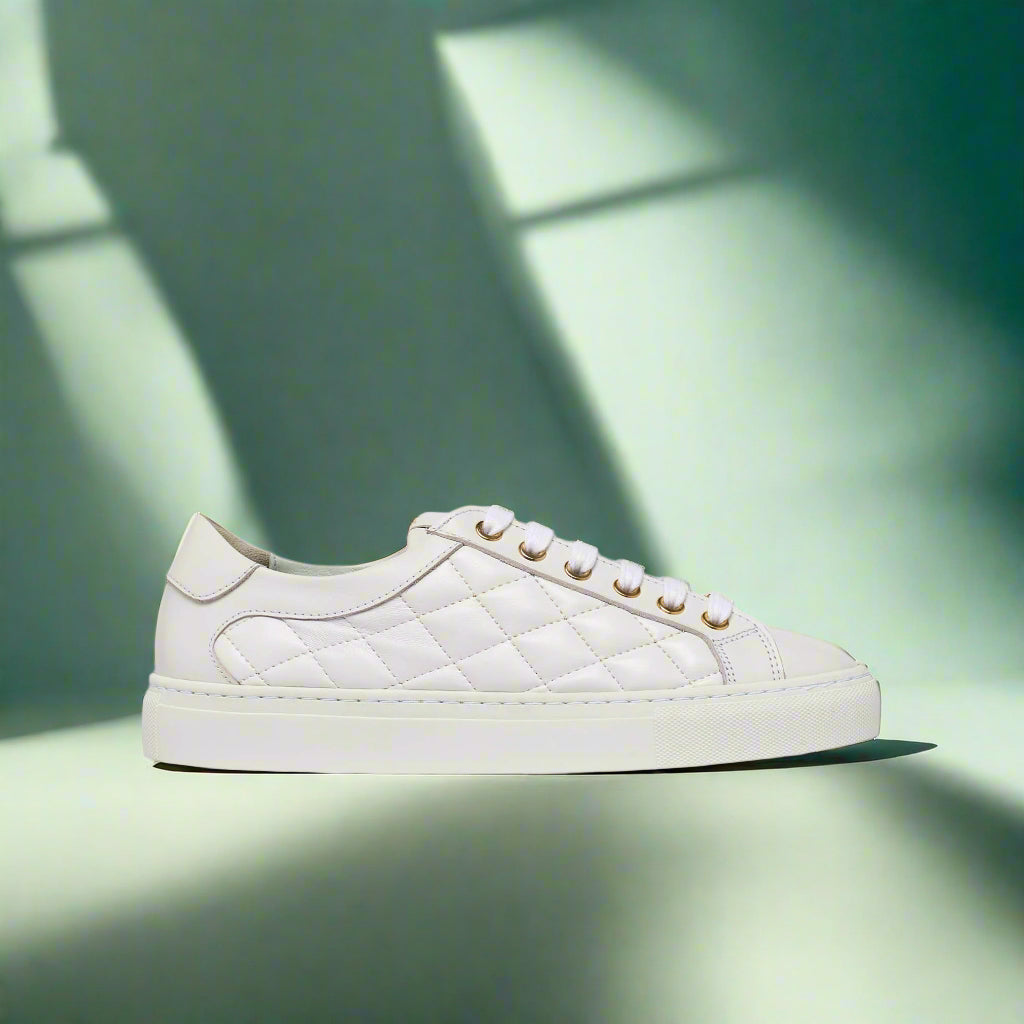 A white quilted sneaker with lace-up closure, displayed against a geometric shadowed background.