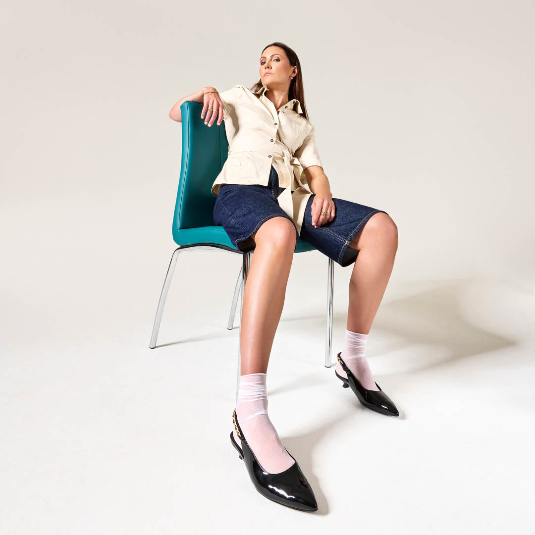 Woman sitting on a teal chair wearing a beige shirt, navy shorts, white socks, and black Thea shoes.