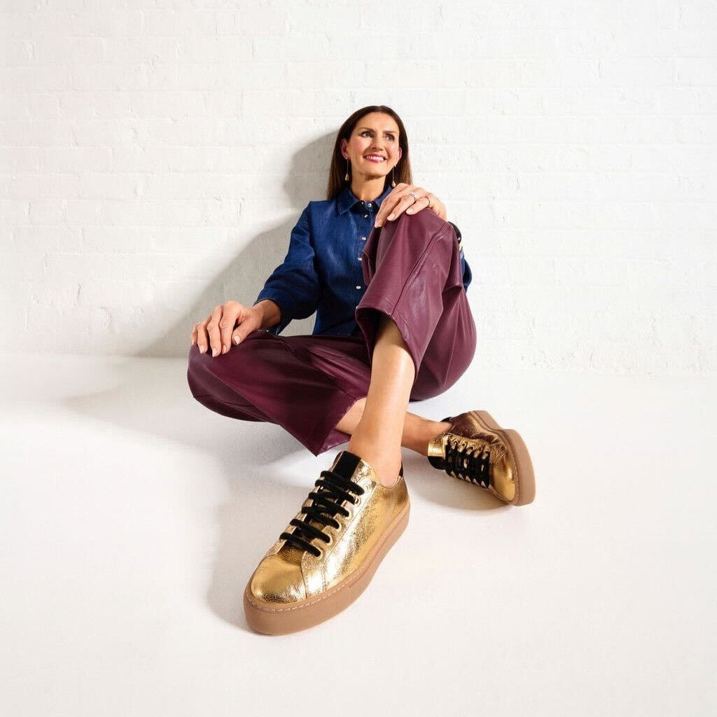 Woman wearing gold sneakers sitting on a white floor.