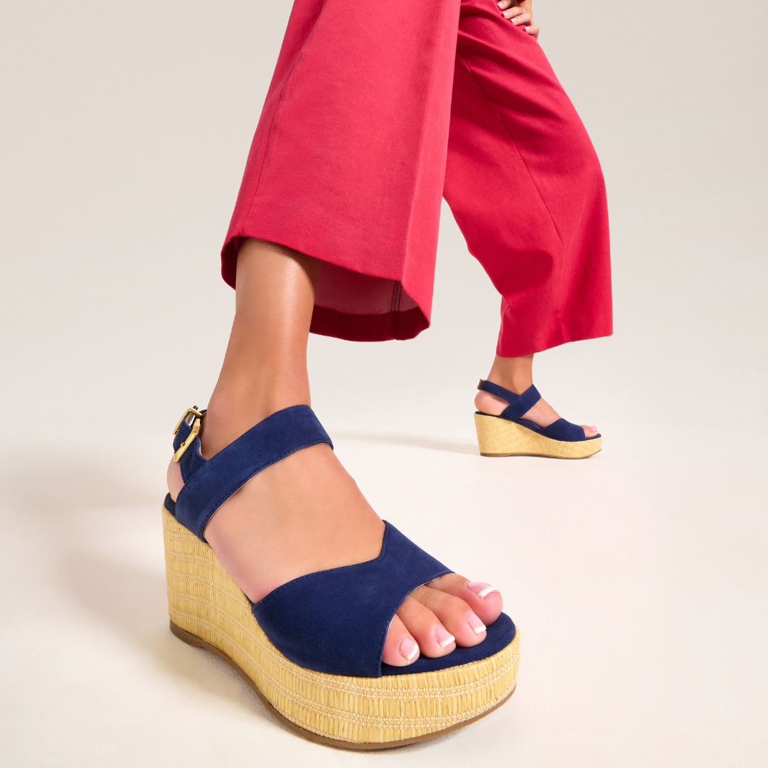 Navy wedge sandals with yellow soles worn by a person in red pants on a light background
