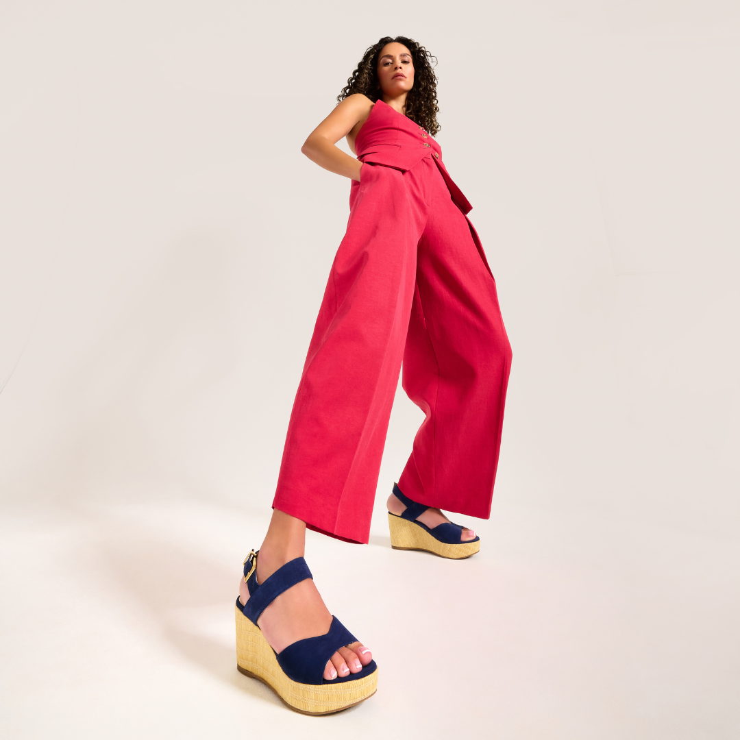 Woman wearing a bright pink jumpsuit with navy sandals on a light background