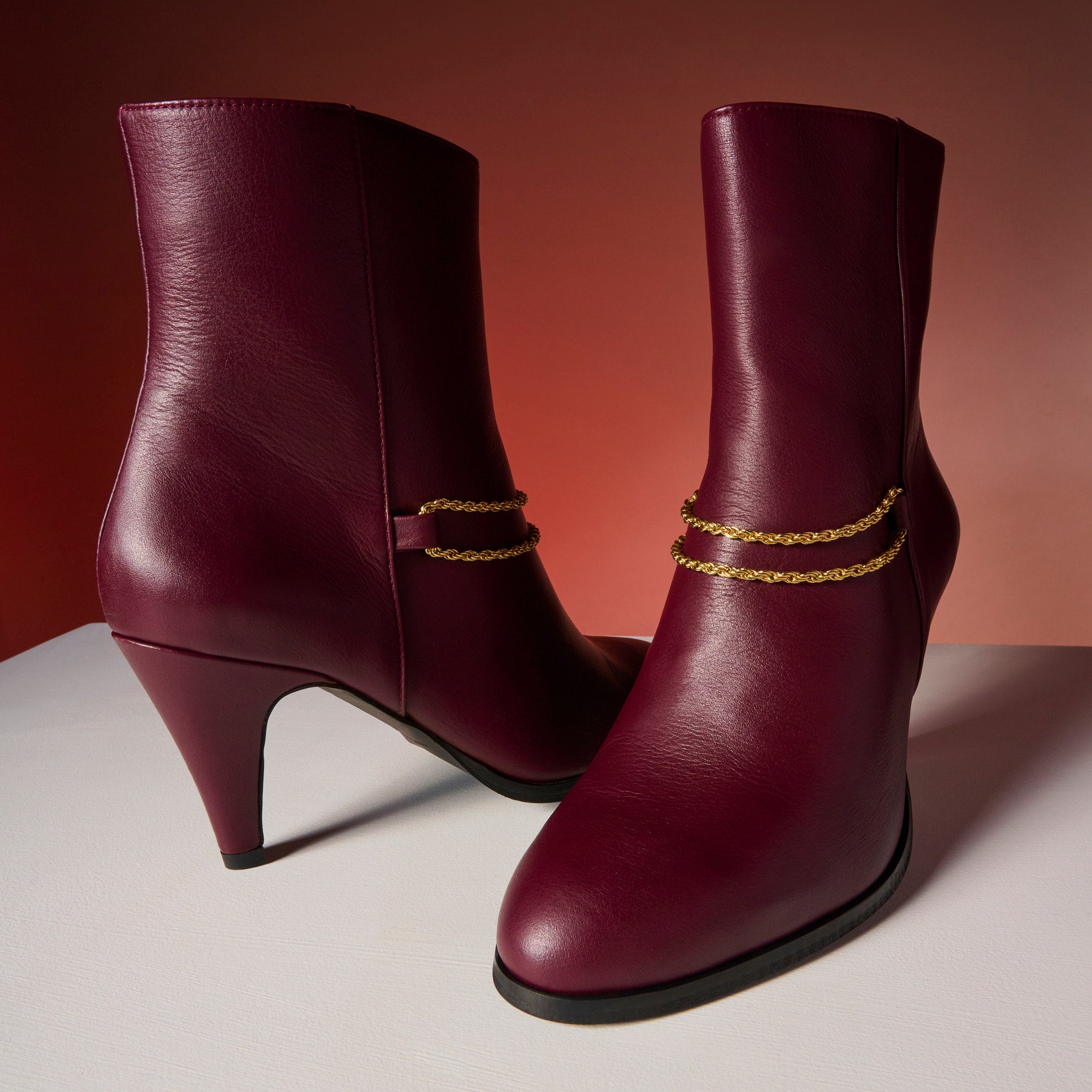 Pair of cherry Maya ankle boots with gold chain details on a gradient background