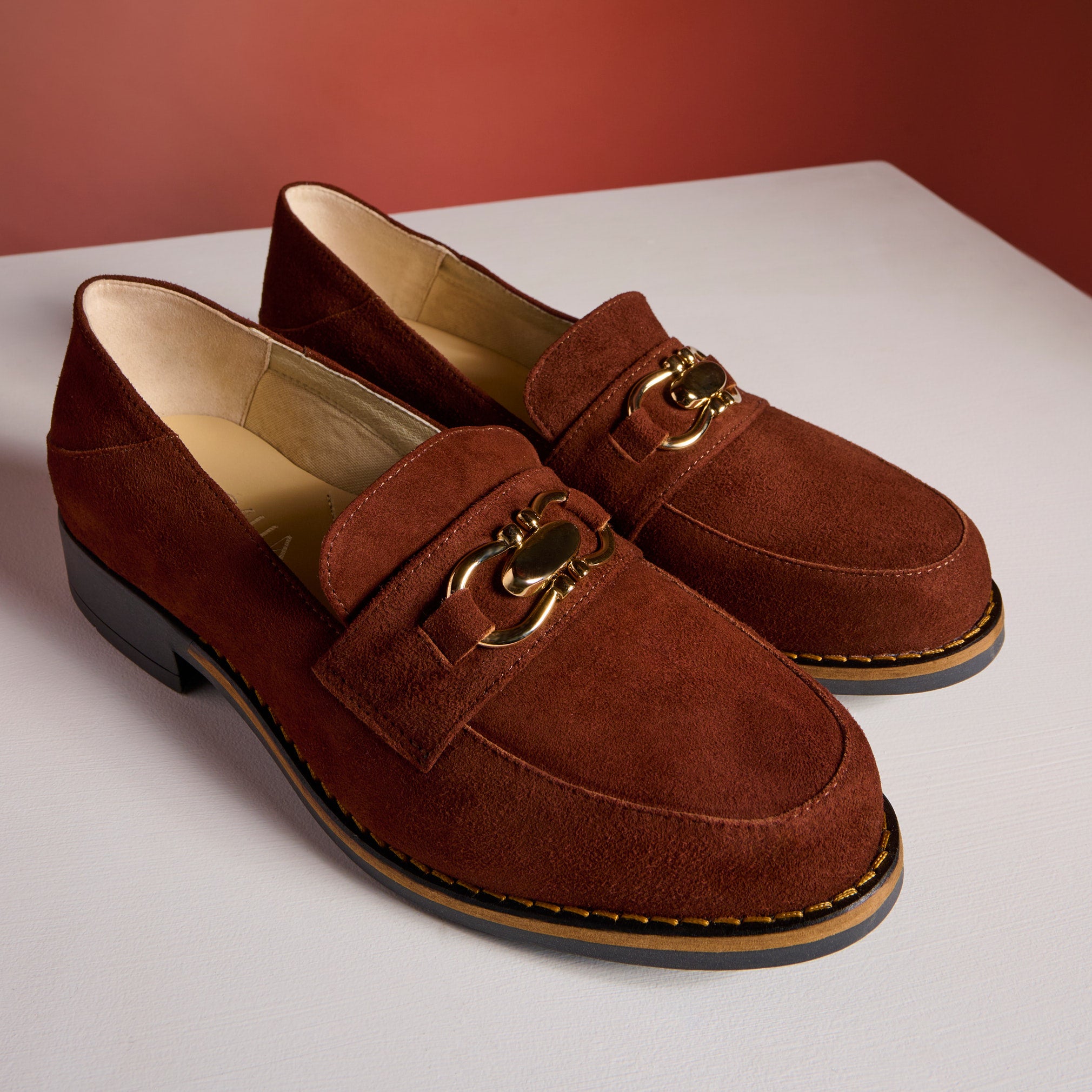 Brown suede loafers with gold chain detail on a white surface.