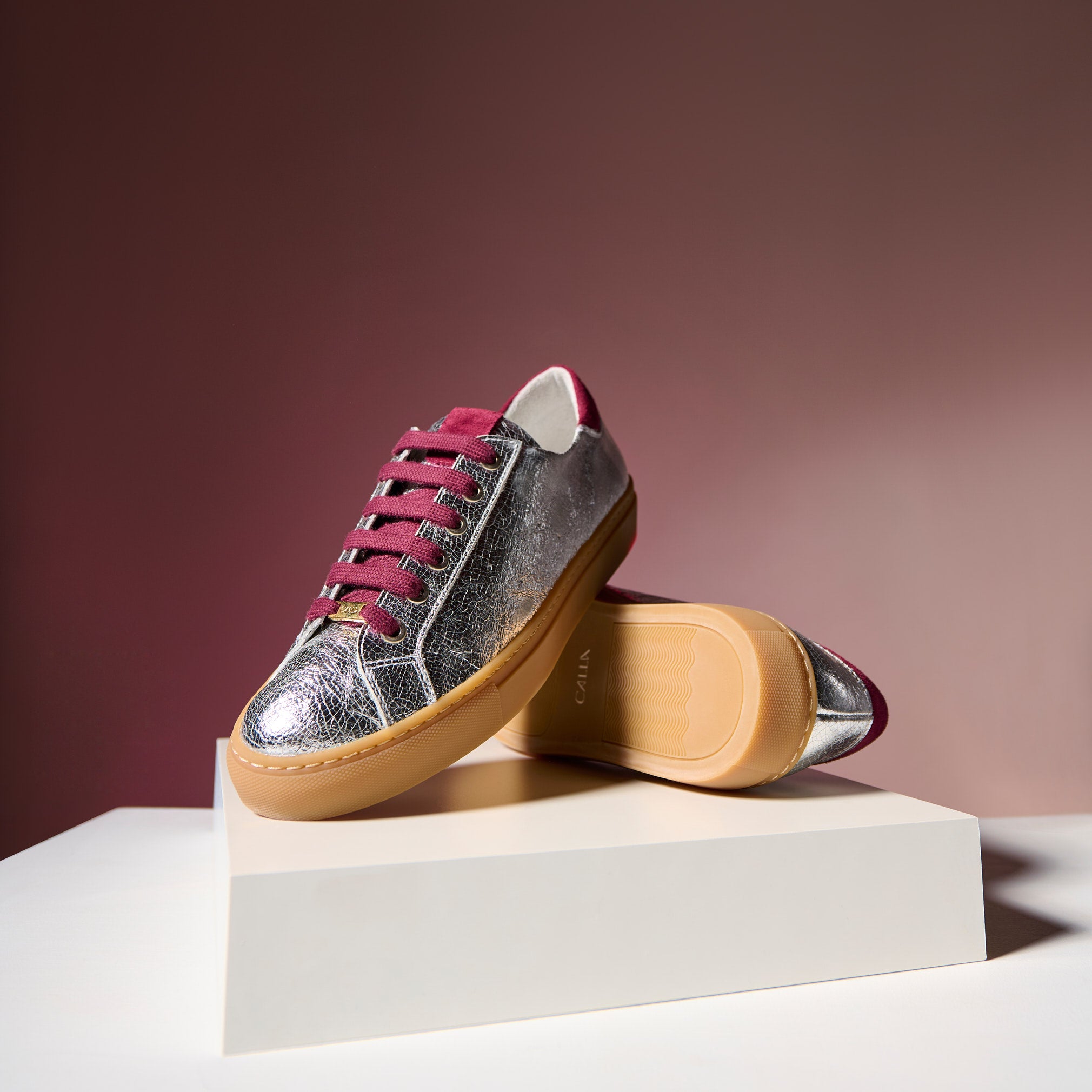 Silver and burgundy sneakers on a beige platform with a maroon background