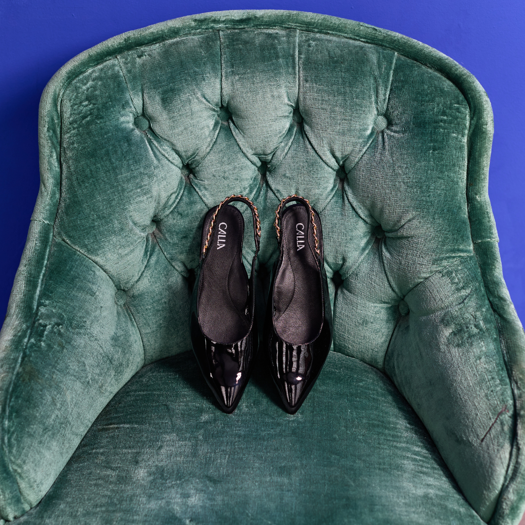 Pair of black Thea shoes on a green velvet chair with a blue background