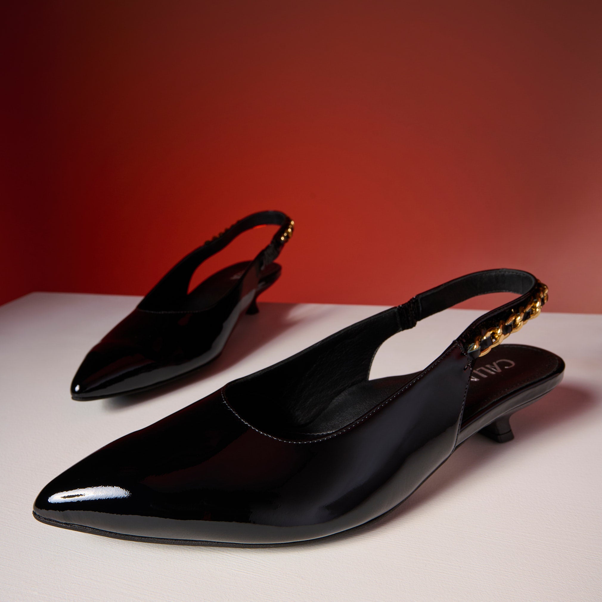 Black patent leather Thea slingback shoes on a white surface with a red background
