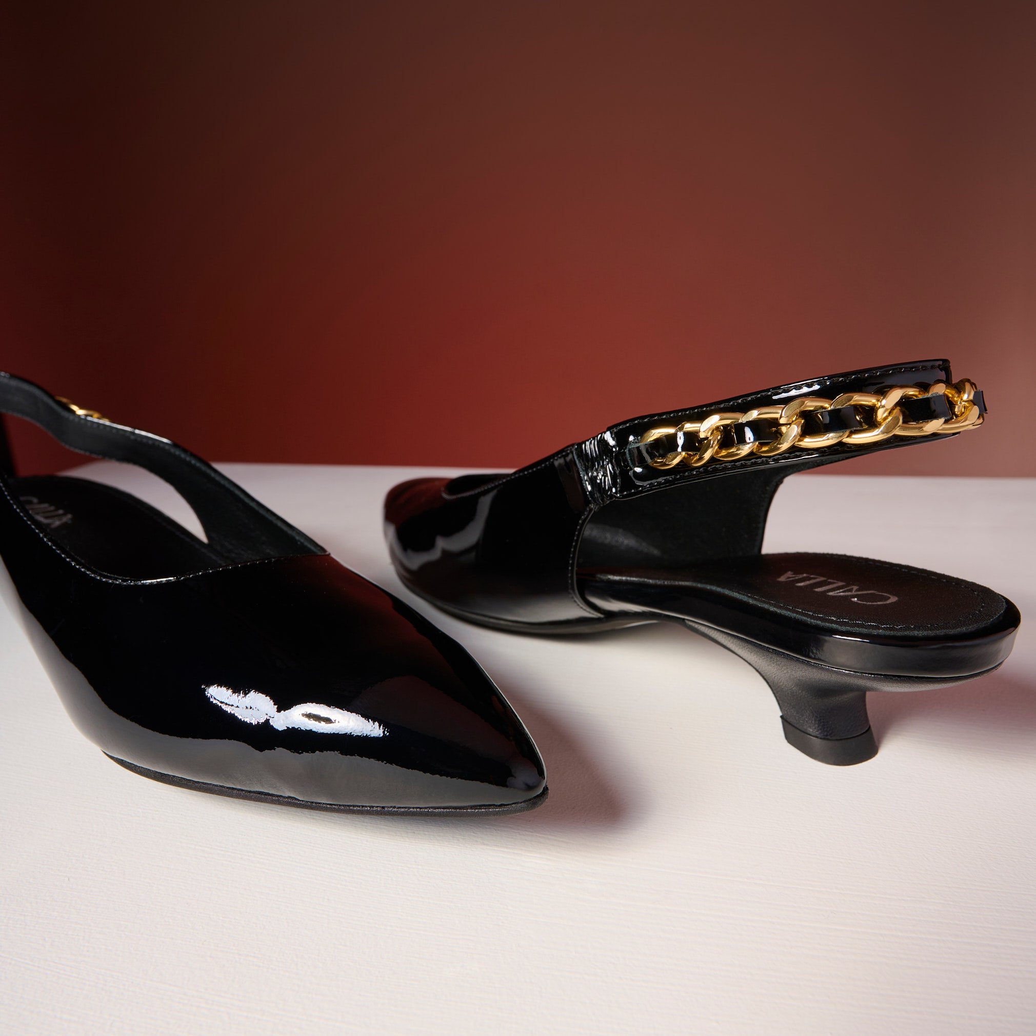 Black Thea patent leather shoes with gold chain detail on a white surface.