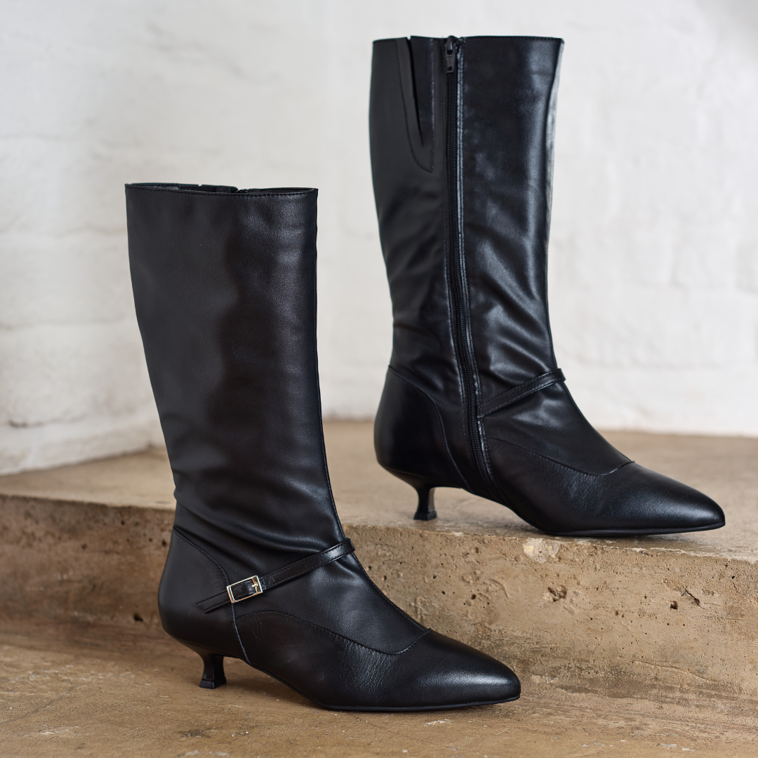 Pair of Veronica black knee-high boots with thin heels on a concrete surface.