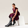 Woman in a purple dress and black knee-high Veronica boots sitting on a white chair against a light gray background.
