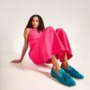 Woman wearing a bright pink jumpsuit and blue loafers sitting on a white background