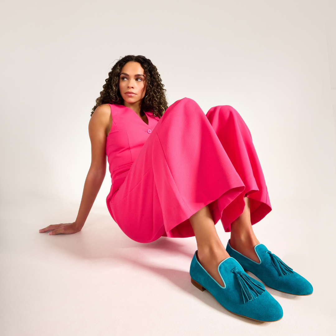 Woman wearing a bright pink jumpsuit and blue loafers sitting on a white background