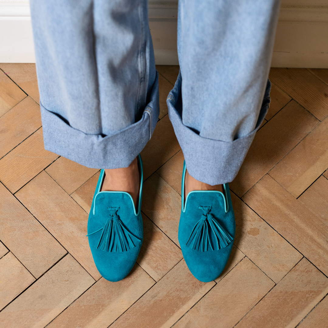 Teal loafers with tassels worn with light blue jeans on a wooden floor.